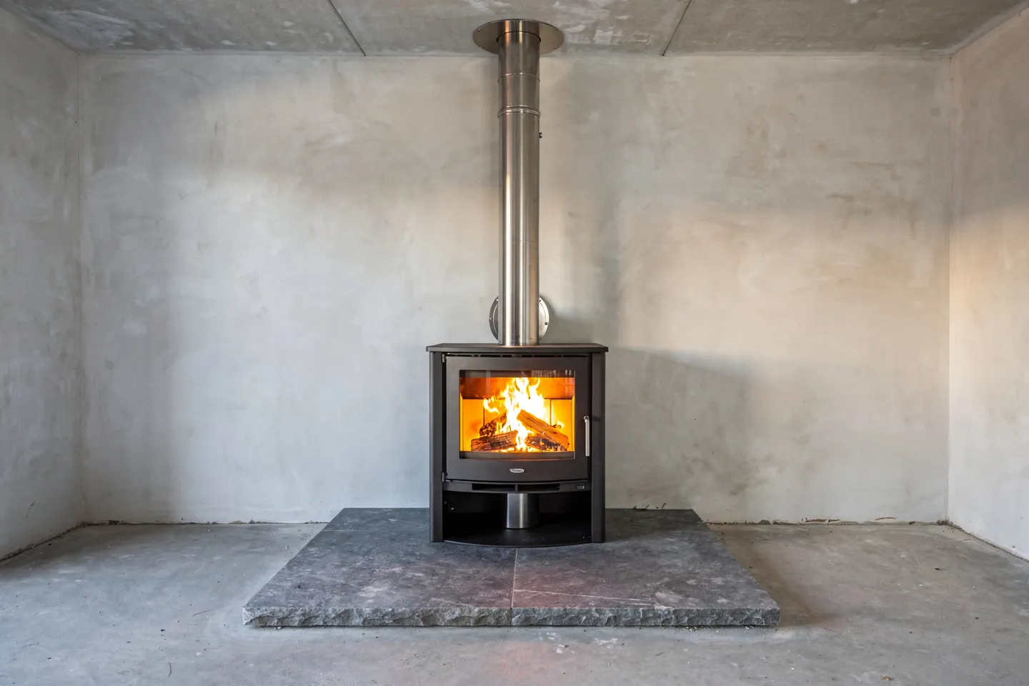 A modern wood-burning stove with a glass door and a small fire on a stone hearth plate in an unfinished room of a new house, a stainless steel flue pipe rising into the ceiling and a small external combustion-air duct entering the stove from behind