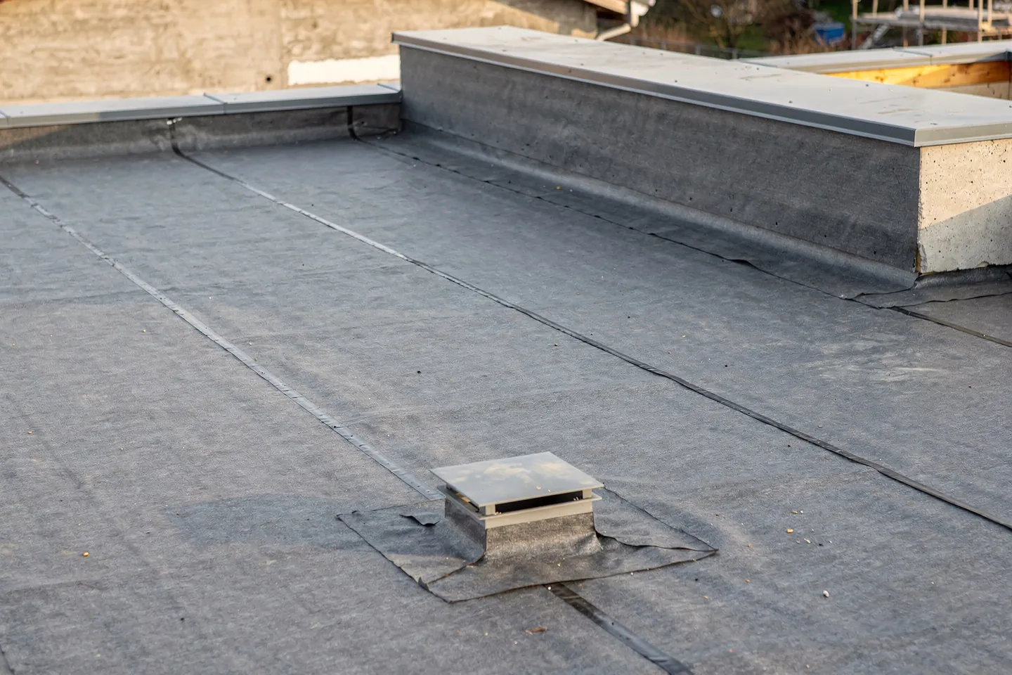 A flat roof on a single-storey house under construction with a grey synthetic membrane laid over tapered insulation boards, a welded seam, a square roof outlet at the low point and a low parapet with metal coping