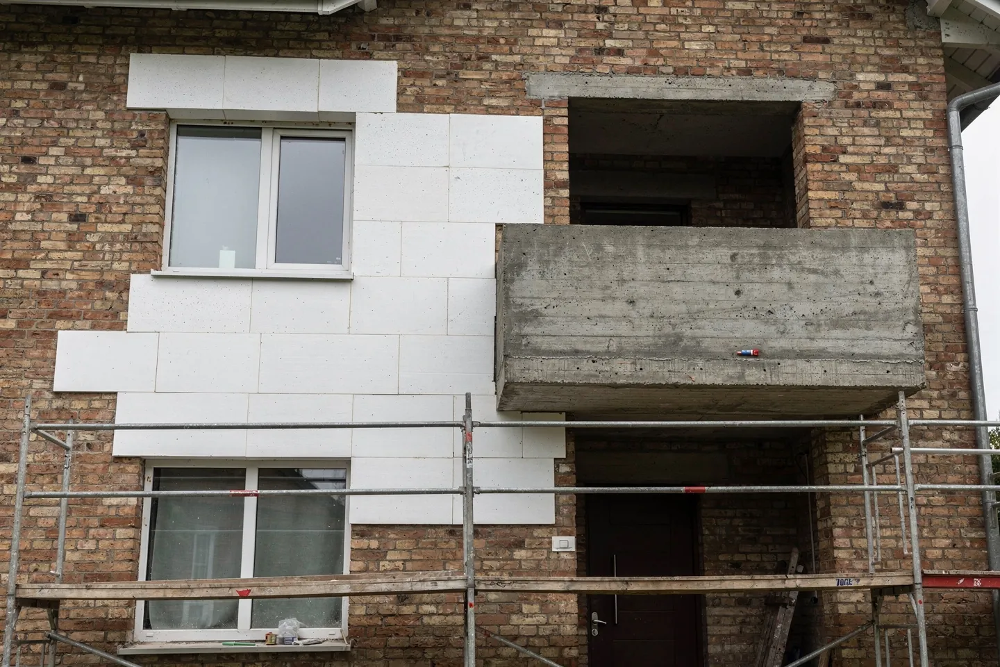 The facade of an old brick family house during renovation with scaffolding in front, new white insulation boards being fixed to the wall, a concrete balcony slab projecting from the wall still uninsulated and insulation cut around a window reveal